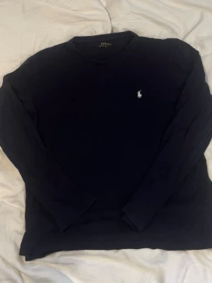 Ralph lauren långarmad - Klassisk mörkblå långärmad tröja från Polo Ralph Lauren med rund halsringning och den ikoniska vita logotypen broderad på bröstet. Perfekt för en stilren och avslappnad look.