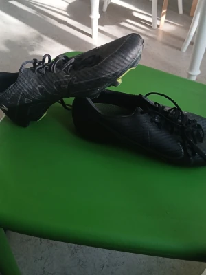 Nike Mercurial svarta fotbollsskor - Säljer ett par svarta Nike Mercurial fotbollsskor med snörning och gula detaljer på sulan. Skorna har låg profil och är tillverkade i syntetmaterial för bra grepp och känsla på planen. Perfekta för dig som vill ha stilrena och snabba skor till matchen.