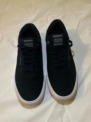 Svarta Jack & Jones skor - Snygga svarta sneakers från Jack & Jones med vita sulor. Skorna har svarta skosnören, diskret logga på sidan och plös, samt ovandel i textilmaterial. Bra för sommaren