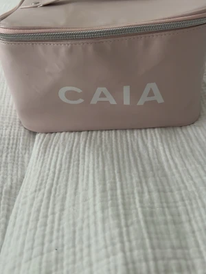 Rosa necessär från CAIA - Snygg och rymlig necessär i ljusrosa färg från CAIA. Väskan har en rektangulär form, dragkedja upptill och ett praktiskt handtag. Tillverkad i ett glansigt syntetmaterial med CAIA-logga framtill. Perfekt för smink eller småprylar.