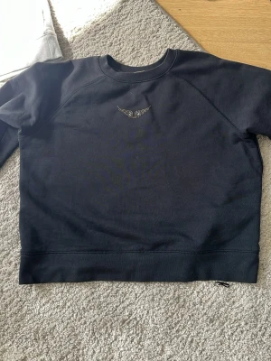Svart sweatshirt från Zadig & Voltaire - Cool svart sweatshirt från Zadig & Voltaire med ett diskret vingmönster i små nitar på bröstet. Tröjan har rund halsringning och är tillverkad i mjukt bomullsmaterial. Kostar i nypris runt 1200kr. Perfekt för en cool och stilren look.