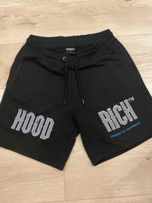 Svarta shorts från Hoodrich. - Svarta shorts från Hoodrich med stora tryck i vitt och grått på framsidan där det står 'HOOD' och 'RICH'. Shorts med snörning i midjan och avslappnad passform. Perfekta för chill dagar och streetwear-stil.