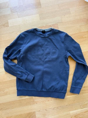 Mörkblå sweatshirt från Hugo Boss - Mörkblå sweatshirt från Hugo Boss med diskret broderad logga på bröstet. Tröjan har rund halsringning, ribbade muddar vid ärmslut och nederkant samt en avslappnad passform. Perfekt för en stilren och enkel look.