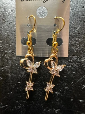 Guldiga örhängen med fjäril och strass - Snygga guldiga örhängen med hängande design, dekorerade med glittrande strass och en söt fjärilsform. Perfekta för att ge din look lite extra bling! Materialet är metall med stenar i klar färg.