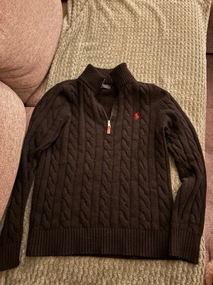 Svart kabelstickad tröja Polo Ralph Lauren - Svart kabelstickad tröja från Polo Ralph Lauren med halv dragkedja och hög krage. Tröjan har ett rött broderat logomärke på bröstet och ribbstickade muddar vid ärmslut och nederkant. Perfekt för lager-på-lager och snygg till jeans.