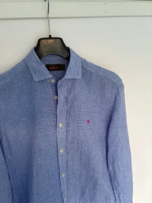 Blå linneskjorta från Morris - S - Snygg blå skjorta i linne från Morris med diskret rutigt mönster och klassisk krage. Skjortan har långa ärmar, knappar framtill och en rosa broderad logga på bröstet. Perfekt för en stilren och avslappnad look.