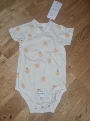Omlott body unisex - Supersöt vit omlott body från Lindex med korta ärmar och tryckknappar. Prydd med gulliga bläckfiskar i beige och grått. Tillverkad i mjuk bomull, perfekt för mysiga nätter och lugna stunder hemma.