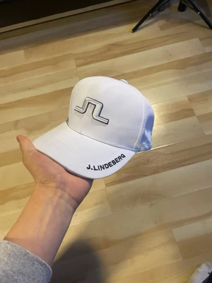 Vit keps från J.Lindeberg - Snygg vit keps från J.Lindeberg med broderad logga framtill och märkesnamn på skärmen. Kepsen har klassisk böjd skärm och justerbar rem baktill. Tillverkad i ett lätt och bekvämt material som passar perfekt till en sportig eller casual stil.