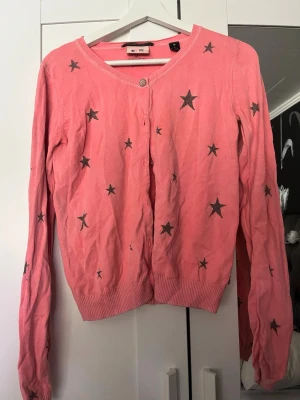 Rosa kofta med stjärnor från Mi Vic - Rosa kofta med svarta stjärnor från Mi Vic. Koftan har rund halsringning, knäppning framtill och ribbade muddar vid ärmslut och nederkant. Perfekt för dig som gillar färg och mönster.