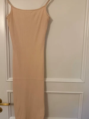 Beige ribbad långklänning - Säljer en beige ribbad långklänning med smala axelband och figurnära passform. Klänningen är ärmlös och har en enkel, stilren design som passar perfekt till sommaren. Materialet känns mjukt och stretchigt för en bekväm känsla.