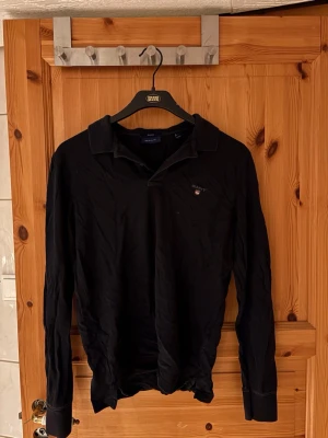 Svart långärmad skjorta från Gant - Klassisk svart långärmad skjorta från Gant med liten logga broderad på bröstet. Skjortan har krage och knappar framtill, tillverkad i mjukt bomullsmaterial. Perfekt för en stilren och enkel look.