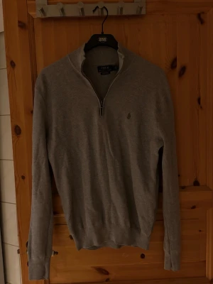 Grå halvzip-tröja från Polo Ralph Lauren - Snygg grå långärmad tröja från Polo Ralph Lauren med halv dragkedja och diskret logga på bröstet. Perfekt för lager-på-lager och enkel att matcha med jeans eller chinos. Klassisk och stilren design.