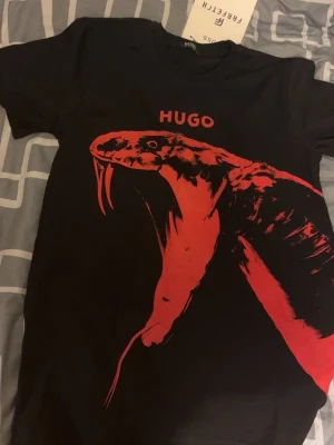 Svart HUGO BOSS t-shirt med röd orm - Cool svart t-shirt från HUGO BOSS med ett stort rött ormmotiv framtill och HUGO-tryck ovanför. T-shirten har klassisk passform och rund hals. Perfekt för dig som vill sticka ut med en unik design.