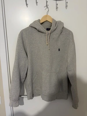 Grå hoodie från Polo Ralph Lauren - Snygg grå hoodie från Polo Ralph Lauren med klassisk broderad logga på bröstet. Hoodien har huva med dragsko och en stor magficka framtill. Tillverkad i mjukt bomullsmaterial som är perfekt för en avslappnad stil.