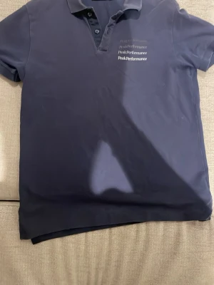 Mörkblå Peak Performance piké - Snygg mörkblå pikétröja från Peak Performance med klassisk krage och knappar framtill. Bröstet pryds av flera Peak Performance-loggor i vitt. Perfekt till jeans eller shorts för en clean look.
