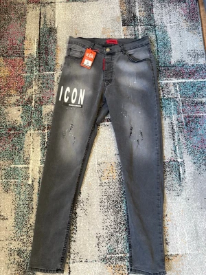 Grå DSQUARED2 ICON jeans - Snygga grå jeans från DSQUARED2 med vit 'ICON'-print på vänster lår och distressed detaljer framtill. Modellen har smal passform, klassiska fem fickor och DSQ2-logga bak i midjan. Tillverkade i stretchigt denim för skön känsla och modern look.