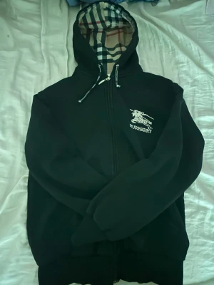 Svart Burberry hoodie med dragkedja - Svart hoodie från Burberry med dragkedja och klassiskt rutigt foder i huvan. Har vit logga och ryttare på bröstet. Justerbar huva med snören och ribbade muddar. Perfekt för en stilren och lyxig streetwear-look.