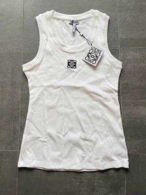 Vit ribbad linnetopp från Loewe - Snygg vit linnetopp från Loewe med ribbad struktur och broderad logga framtill. Modellen är ärmlös med rund halsringning och tillverkad i mjuk bomull. Perfekt att styla med jeans eller kjol för en clean look.