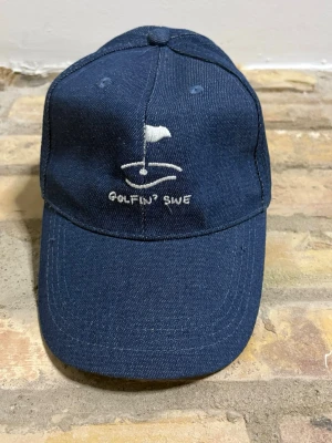 Mörkblå keps GolfinSWE  - Snygg mörkblå keps från Golfing SWE med broderad vit golf-flagga och text framtill. Klassisk böjd skärm och justerbar baksida. Tillverkad i denimliknande material som ger en cool och avslappnad look.