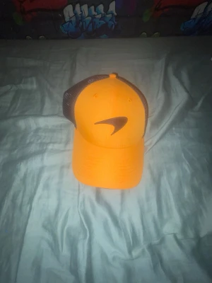 Orange McLaren keps från New Era - Snygg orange keps från McLaren Formula 1 Team i samarbete med New Era. Kepsen har svart mesh på sidorna och bak, böjd skärm och justerbar passform. Perfekt för dig som gillar racing och streetstyle.