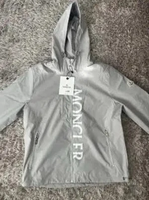 Grå vindjacka från Moncler - Snygg grå vindjacka från Moncler med huva och dragkedja framtill. Jackan har stora vita Moncler-bokstäver längs dragkedjan och två fickor med dragkedja. Perfekt för blåsiga dagar och streetwear-stil. Storlek XL men passar L,M