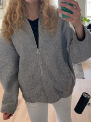 Grå teddyjacka med dragkedja - Mysig grå teddyjacka med dragkedja framtill och avslappnad passform. Jackan har långa ärmar och är tillverkad i fluffigt teddy-material som ger en cool och trendig look. Perfekt för att hålla värmen under kyliga dagar.
