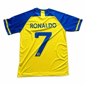 Cristiano Ronaldo #7 Al-Nassr Home Jersey 22/23 - Säljer en officiell Al-Nassr hemmatröja från säsongen 2022/2023 med Cristiano Ronaldo #7 på ryggen. Tröjan är i den klassiska gul-blå designen med alla detaljer intakta.  Märke: Duneus Sportswear (officiell leverantör för säsongen).  Storlek: Large (L).  Skick: Mycket gott skick. Trycket på ryggen är tydligt och utan sprickor. Klubbemblem och sponsorer (Shurfah) sitter perfekt.  Detaljer: Inkluderar autentiska detaljer som "D-TECH" och säsongens officiella märkning nertill. Skickas snabbt! 