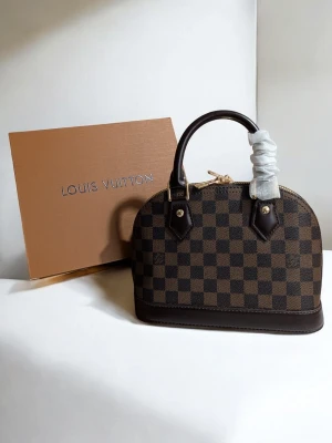 Iouis Vuitton Alma BB. - En tidlös famn av elegans Denna vackra skapelse vilar med en naturlig värdighet, klädd i ett djupt och harmoniskt schackrutigt mönster i varma toner av choklad och ebenholts. Dess mjuka, rundade former viskar om en klassisk skönhet som aldrig går ur tiden, en trofast följeslagare som bär dina hemligheter med stil.