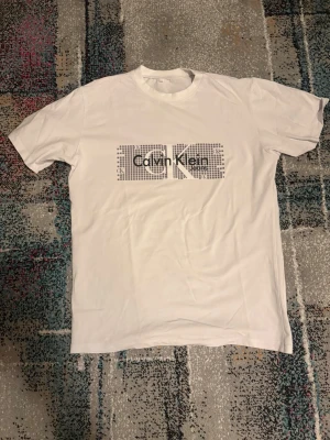 Vit Calvin Klein t-shirt med tryck - Vit t-shirt från Calvin Klein Jeans med ett stort grafiskt tryck i svart och grått på bröstet. Klassisk rund hals och korta ärmar. Perfekt för en clean och stilren look.