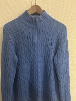 OANVÄND!!Blå kabelstickad tröja Polo Ralph Lauren - Kabelstickad blå tröja från Polo Ralph Lauren med halv dragkedja och hög krage. Tröjan har det klassiska broderade logotypen på bröstet och ribbade muddar vid ärmslut och nederkant. Perfekt för lager på lager och snygg till jeans. PRIS KAN DISSKUTERAS VID INTRESSE OCH SNABBT KÖP