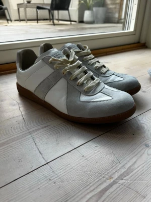 Vita sneakers med grå detaljer - Snygga sneakers i vitt läder med grå mockadetaljer och bruna gummisulor. Klassisk låg modell med vita snören och stilren design som passar till det mesta. Perfekta för dig som gillar en clean och tidlös look.