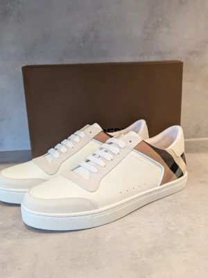 Burberry vita sneakers med rutigt mönster - Stilrena vita sneakers från Burberry med klassiskt beige, svart och rött rutigt mönster på hälen. Skorna har vita snören, diskreta perforeringar på sidan och detaljer i mocka. Perfekta för dig som vill ha en clean look med lyxig touch.