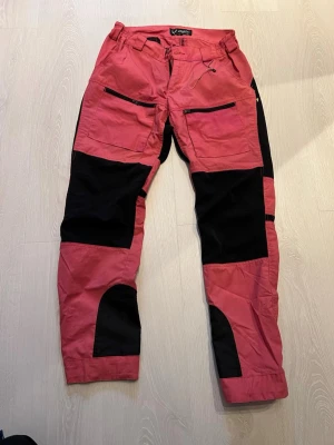 Rosa och svarta cargobyxor Jägarliv - Snygga rosa och svarta cargobyxor från Jägarliv med flera praktiska fickor och dragkedjor. Byxorna har förstärkta partier på knän och ben, perfekt för uteliv och äventyr. Tillverkade i slitstarkt material som passar för outdoor och friluftsliv.