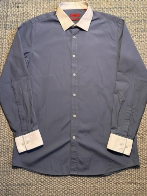Blå skjorta med vit krage från Hugo - Snygg blå skjorta från Boss med vit kontrastkrage och vita manschetter. Skjortan har slim fit passform, långa ärmar och klassiska knappar framtill. Perfekt för dig som gillar stilrena detaljer och modern design.