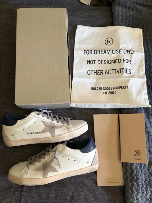 Golden Goose vita sneakers med stjärna - Golden Goose sneakers i vitt läder med grå mockastjärna på sidan och grå snörning. Hälpartiet är mörkblått och sulan har en vintage-look. Klassisk låg modell med GGDB/SSTAR-tryck på sidan. Kommer med originalkartong och dustbag.