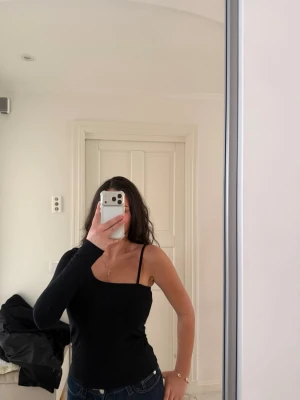 Svart oneshoulder topp - Trendig svart oneshoulder topp med en lång ärm och en axelbandslös sida. Sitter tight och har en stilren look som passar perfekt till jeans eller kjol. Enkel och snygg design som ger en cool vibe.