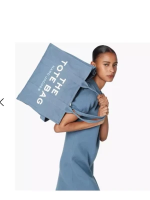 Blå totebag Marc Jacobs - Snygg totebag från Marc Jacobs i blå canvas med vita tryckta bokstäver 'THE TOTE BAG'. Väskan har dubbla handtag och rymlig design, perfekt för att bära allt du behöver. Klassisk och trendig accessoar för dig som gillar stilrena detaljer. Lite solblekt på tex axelbanden men inget slitet på väskan💕 haft den som skolväska och man får verkligen plats med ALLT💕 får inte mycket användning för den längre då jag köpt en ny väska💕nypris 3500k köpt på NK💕