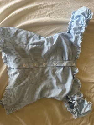 Blåvit randig blus med volang - Söt blus i blåvitt randigt tyg med volangdetaljer längs ärmar och axlar. Blusen har knappar framtill och är tillverkad i ett lätt bomullsliknande material. Perfekt för en fräsch och somrig look.