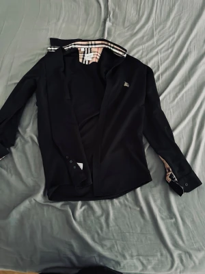 Svart skjorta från Burberry - Stilren svart skjorta från Burberry med klassiskt rutigt mönster på insidan av kragen och manschetterna. Skjortan har långa ärmar, knappar framtill och en liten logga på bröstet. Perfekt för dig som gillar exklusiva detaljer.