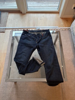Ralph Lauren selvedge jeans - Säljer ett par mörkblå jeans från Ralph Lauren med raka ben och snygga detaljer. Baksidan har en stor broderad patch med guldfärgade och röda inslag ovanför ena bakfickan. Jeansen har knäppning fram och klassisk femficksdesign.
