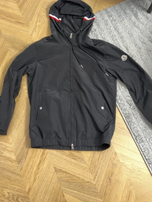 Svart vindjacka från Moncler - Svart vindjacka från Moncler med huva och dragkedja framtill. Jackan har två fickor med tryckknappar och Moncler-logga på ärmen. Röd och vit detalj på huvan. Lätt och smidig modell i syntetmaterial.