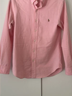 Rosa skjorta från Ralph Lauren - Klassisk rosa skjorta från Ralph Lauren med button-down krage och broderad logga på bröstet. Skjortan har långa ärmar, knappar framtill och är tillverkad i mjuk bomull. Perfekt för en stilren och avslappnad look nu till sommaren 🍻☀️