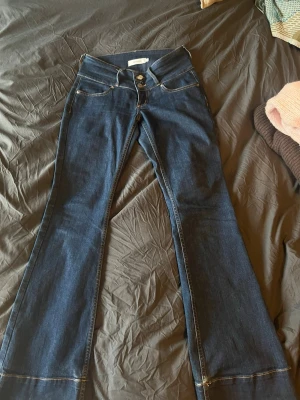 Mörkblå bootcut jeans Junkyard - Snygga mörkblå bootcut jeans från Junkyard med hög midja och dubbla knappar framtill. Klassisk femficksmodell med kontrastsömmar och stretchigt denimtyg som sitter skönt. Perfekta för dig som gillar retrovibbar och vill ha ett par jeans som sticker ut. Lite slitna längst ner där bak, men annars bra skick🥰