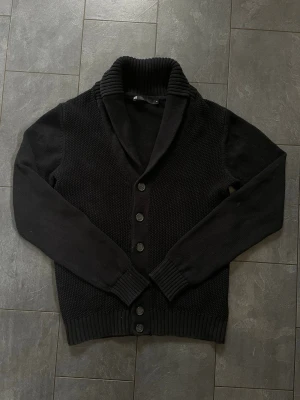 Svart cardigan från WE - Säljer en svart stickad cardigan från WE och fem knappar framtill. Cardiganen har ribbstickade muddar och nederkant samt en grovstickad struktur på kroppen. Perfekt för dig som gillar stilrena och klassiska plagg.