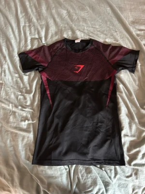 Svart och vinröd träningströja Gymshark - Snygg svart och vinröd träningströja från Gymshark med korta ärmar. Tröjan har ett sportigt mönster över bröst och rygg samt Gymshark-logga framtill. Tillverkad i ett stretchigt och snabbtorkande material som passar perfekt till gymmet eller löpning.