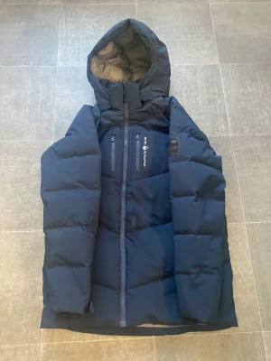Mörkblå Sail Racing Antarctica jacka  - Snygg mörkblå dunjacka från Sail Antarctica Expedition med hög krage, huva och flera dragkedjefickor. Jackan har en cool patch på bröstet och är perfekt för kalla vinterdagar. Tjockt vadderad för extra värme och stil. Använd en vinter endast. Den passar till 170cm-185cm!