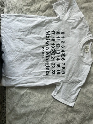 Maison Margiela vit t-shirt med tryck - Vit t-shirt från Maison Margiela med ikoniskt svart tryck framtill med siffror och logga. Klassisk passform med rund hals och korta ärmar. Tillverkad i mjuk bomull som känns skön mot huden. Perfekt för dig som gillar stilrena och trendiga plagg.