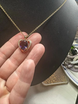 Halsband med hjärthänge och lila sten - Snyggt halsband med tunn guldfärgad kedja och ett hjärtformat hänge. Hänget har en stor lila sten i mitten, omgiven av små klara stenar som ger extra bling. Perfekt accessoar för dig som gillar unika smycken med elegant touch.