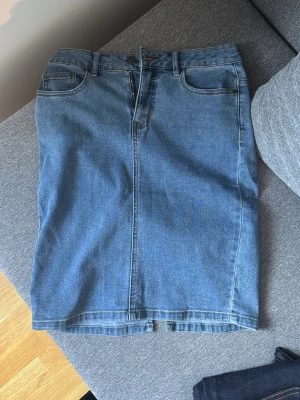 Blå jeanskjol med slits bak - Säljer en klassisk blå jeanskjol i rak modell med fem fickor, bälteshällor och slits bak. Kjolen har dragkedja och knapp framtill och är tillverkad i ett tåligt denimtyg. Perfekt att styla med sneakers eller boots för en avslappnad look.