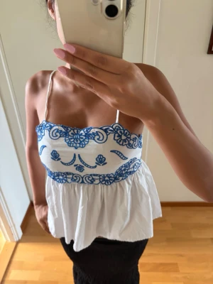 Vit och blå broderad linnetopp - Söt vit linnetopp med tunna axelband och blått broderat blommönster upptill och i midjan. Toppen har en luftig och flowy passform, perfekt för varma dagar. Materialet känns lätt och toppen är ärmlös med en feminin touch.